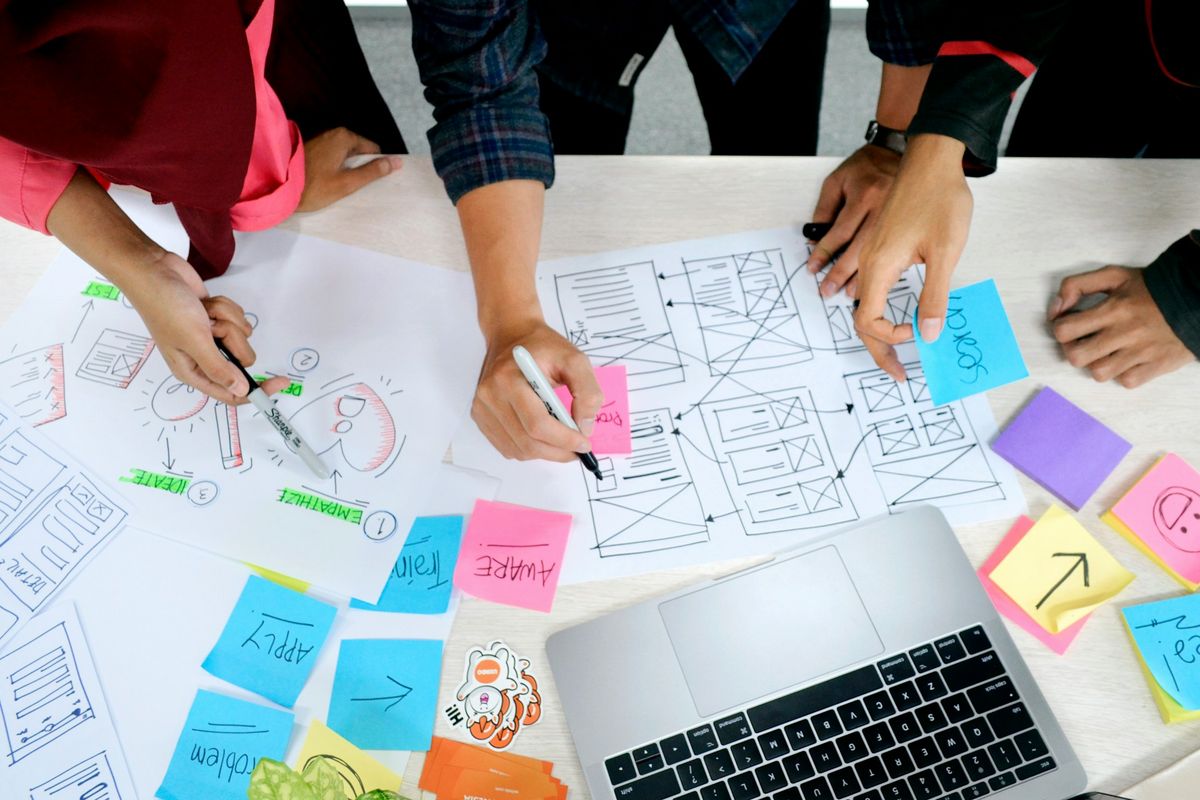 Overhead view of a desk with partial view of a laptop as well as various paper mockups of website and application pages. There are several colourful sticky notes on them and the hands of 3 people hovering above the papers, with pens in hand.